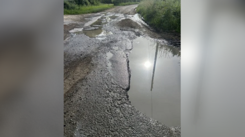 A damaged road covered with large potholes filled with water and grass verges on either side.