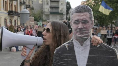 A woman wearing sunglasses makes an announcement over a PA system as she holds a cutout of a man. Behind are other people in the streets, with one person holding a Ukrainian flag