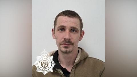 A police custody image of a man with short dark hair and short facial hair, wearing a green hoodie and stood in front of a light-coloured wall. There is a West Mercia Police logo in the bottom left corner.