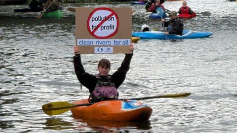 A woman sat in a kayak holding a sign reading "clean rivers for all"