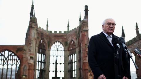 German president Frank-Walter Steinmeier stands behind microphones in front of the ruins of Coventry Cathedral. He is wearing glasses, and a grey tie, white shirt and black jacket. 