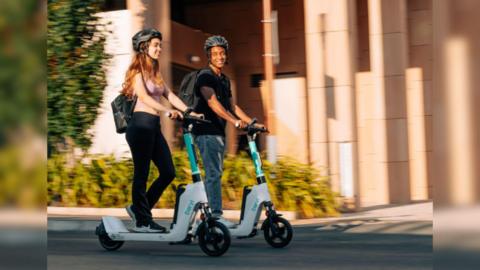 Two people riding on white and green e-scooters on a road