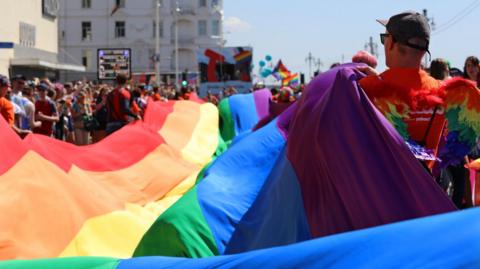 A man holding one side of a large rainbow flag. The person is carrying the flag in a parade through a city street lined with people.