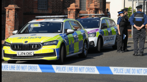 Police cars on a street in front of a house with police tape in the foreground and uniformed officers in background