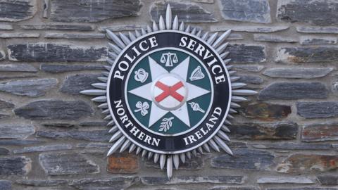 The PSNI badge on a stone wall. The badge is a Bath star with lettering around the inside reading "police service of Northern Ireland". In the centre is a red and white St Patrick's Saltire.