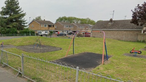 Google shot of Fern Park play area on Pinchbeck Road in Spalding. It is a lawned area with various apparatus, sone of which appears to be out of order. 