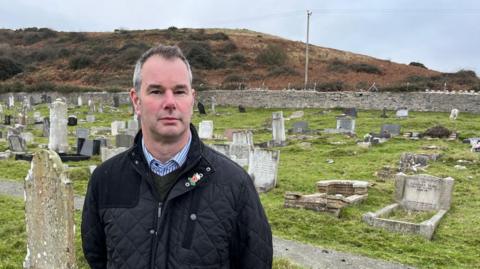 Adrian Hughes standing in a cemetery looking at the camera. He is wearing a black coat and on the left there is a poppy badge. There are headstones behind him.