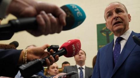 Michéal Martin, who has thinning light brown hair, and is wearing a navy suit talks to reporters who are holding put microphones towards him