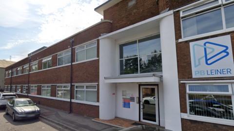 The PB Leiner building from Google Street View. There is a sign with the company name and logo, a styled letter p and letter b. the building is two storey brick with wide double windows on both floors, extending for twelve windows on each floor from a single windowed door entrance. 