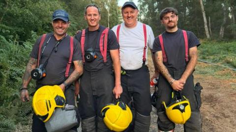 Four firefighters stood in a forest smiling at the camera. They have their protective black trousers on with red ties that go over their shoulders, and they are carrying yellow helmets. Three of them have black T-shirts on, and the other is wearing a white one. Two of them are wearing caps.