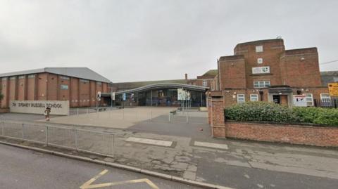 Google StreetView image of the secondary school site for Sydney Russell School, a large brick building with a stone and metal sign to the left of the entrance