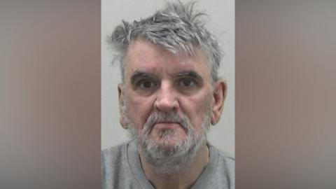 Mugshot of David Elliot. He has thick scruffy grey hair and an untidy grey beard