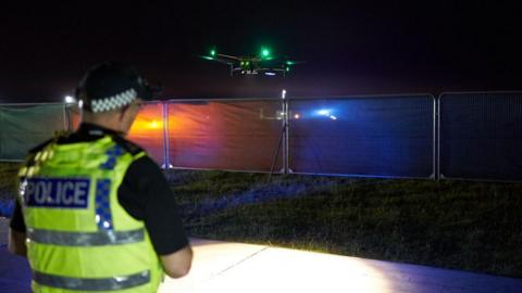 Image shows a police officer in a high-vis vest and headtorch operating a drone at night over a field.