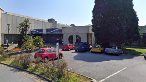 A photo of the outside of the vaccination centre. It is a big building with a blue entrance sign above the door. There is a car park outside with lots of cars in it. There is also plenty of greenery and a big tree outside. 