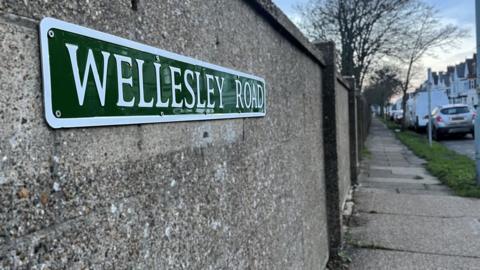 A road sign saying Wellesley Road screwed to a wall. Parts of the road can be seen in the background