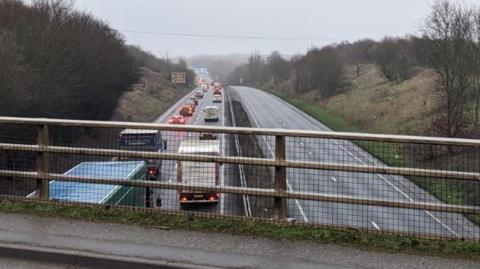 A view of the A47 from a bridge running above it. The left hand carriageway is filled with cars travelling away from the camera in long queues. The right hand carriageway is empty due to the closure.