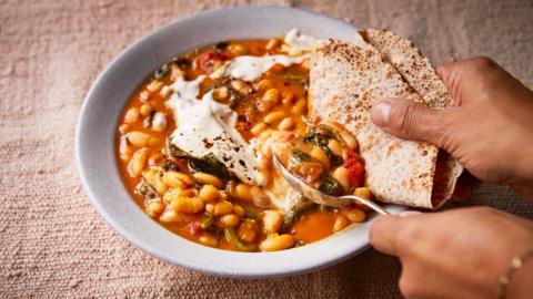 A white bowl of bean curry with a hand dipping flatbread into it