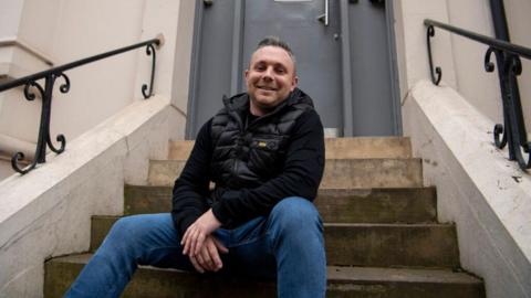 A man with grey hair is sitting on the front steps of a white building and is smiling at the camera. He is wearing a black top, black puffer gilet and blue jeans.