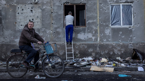 A man stands on a ladder to cover a broken window damaged by a Russian missile strike in the town of Dobropillia, Donetsk region. Another man cycles past the building. The pavement outside the building is strewn with debris and shrapnel.