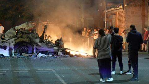 A crowd looks on as a double-decker bus is engulfed by flames as disorder breaks out in Leeds