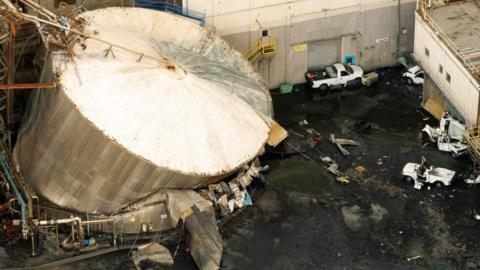 A drone view from the air of the ruptured chemical tank shows a huge white and brown cylindrical tank pitched onto its right hand side, with a black substance flooded onto the ground. Three white vehicles can be seen abandoned within the walls of the facility