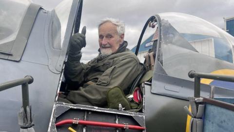 A man sat in a plane cockpit holding his thumb up to the camera