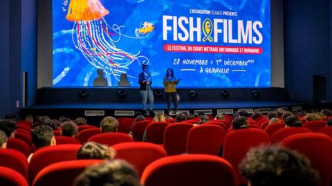 Two people talk to a room full of cinema goers sat in red chairs, with a blue screen and Fish and Films logo projected onto the screen behind.