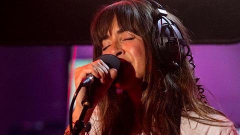 Abi Harding of The Zutons wears a white t-shirt and headphones and holds her saxophone as she sings in a purple and black radio studio