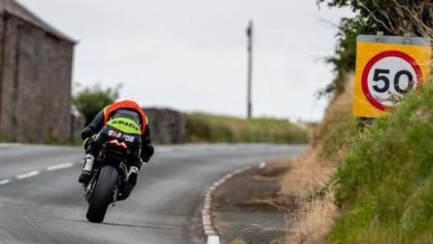 A rider in black with an orange bib negotiates a bend. There is a 50mph road sign above the grass bank on the right.
