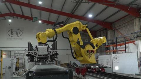 A large yellow-coloured robot on a track facing a conveyor belt. It is in the shape of a large arm, and on the end has a rectangular-shaped contraption which is used to manipulate the components.