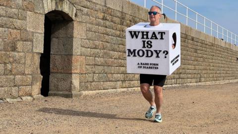 A man wearing a sugar cube costume which says "WHAT IS MODY" and "A RARE FORM OF DIABETES" on the front of it. He is wearing black cycle shorts. He is running on brown gravel. He is wearing sunglasses. Behind his is a brown wall. The sky is blue.