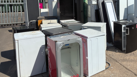 About 10 appliances, with around half of them white, are near each other outside. Railings are in the background on the left.