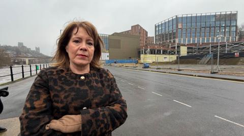 MP Mary Kelly Foy standing across the road from the almost finished Milburngate development. Foy is looking at the camera with a serious expression. She has shoulder-length brown hair and is wearing a leopard print-type coat with golden buttons. To the left is the river Wear and to her right is the development. It is made of a main building with window walls that looks finished. In front of it is heras fencing around the perimeter.