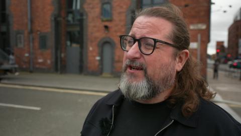 Barry Dallman, who has a beard, a ponytail and is wearing glasses and a black zip-up top, is standing outside a former warehouse building in Liverpool's Baltic Triangle area