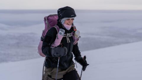 A woman in black arctic gear with black hiking poles is smiling. The hood is up and she has a pink neck scarf around her chin. There is a pink rucksack on her back with water bottles on the straps. Beyond her is a vast expanse of white snow and ice.