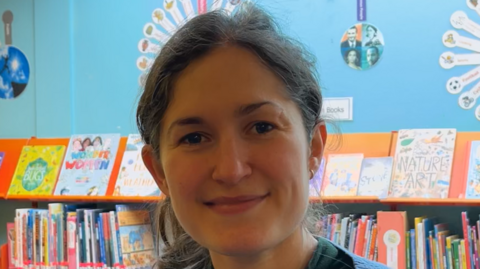 Lizzie Edwards smiles at the camera inside a library. Books can be seen stacked on a shelf behind her. She has brown hair that is tied back behind her head.