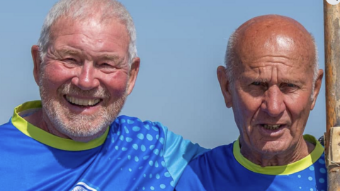 Glenn Powell (left) and Ross Fisher. They are two 78-year-old men wearing blue T-shirts. Glenn has grey stubble and hair while Ross is clean shaven and bald