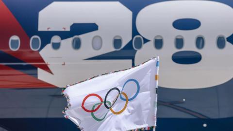 The Olympic flag is flown in front of a plane with LA 2028 branding