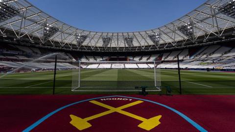 A wide-angle view from behind a football goal net looking into the empty London Stadium under a clear blue sky, featuring the West Ham United crossed hammers logo painted in yellow on the ground in the foreground.