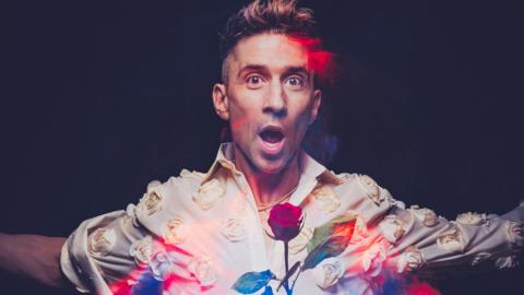 An artistic image of comedian Russell Kane - a man with short dark hair, wearing a white shirt with white flowers on it. A rose is also in the foreground. His mouth is wide open and his arms are open.