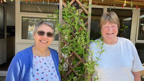 Debi Rice stands on the left of the photo in a floral t-shirt and blue cardigan. Lucia Dedear is on the right of the photo in a grey t-shirt. In between them is a garden trellis and behind them is a house.