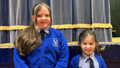 Isla hsa long brown hair, which is draped over her shoulder. She is wearing a blue school uniform and is standing near a stage with a blue curtain. Hannah, a P3 pupil, is standing beside Isla. She has brown hair and is wearing a light blue shirt and tie with a blue cardigan. 