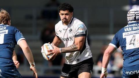 Theo McFarland (centre) stands between two Sale players holding a rugby ball in two hands during a game for Saracens
