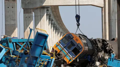 A damaged train on its side surrounded by twisted metal with elevated train tracks in the background