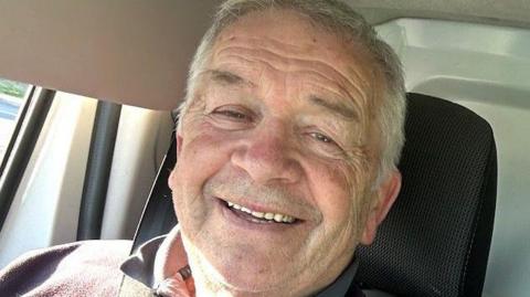 Stuart is smiling at the camera whilst sat in a car. He has short grey hair and is wearing a grey top