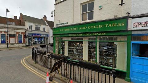 The jewellery shop. Items are displayed in the window and the shop is painted green. There are traffic cones and a sign in the road.