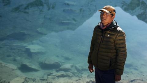 Peruvian farmer Saul Luciano Lliuya poses for a photo in front of Lake Palcacocha. He is wearing a baseball cap and behind him the clear waters of the lake can be seen. 