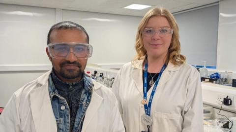 A man stands on the left wearing a white lab coat over a denim jacket with safety goggles over his glasses. He has short dark hair and dark facial hair. To his right, a woman stands wearing a white lab coat done up with a blue staff lanyard and necklace on. She has shoulder length curled blonde hair, behind then is a lab desk.