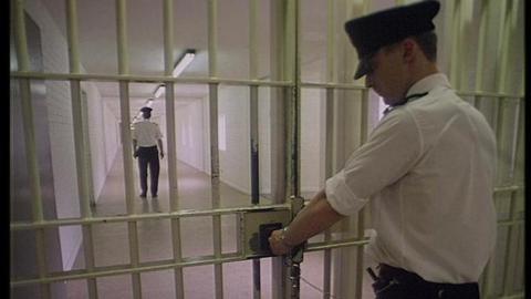 A prison officer locks a pair of metal gates at the end of a corridor. Further away is another prison officer. They are wearing white shirts, black trousers and have peaked black caps