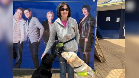 Joy Drury with dark hair and wearing a grey top with two dogs standing outside Marco's Cafe on Barry Island